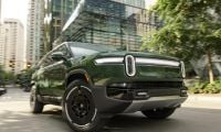 2023 Rivian R1S electric SUV in forest green, shown at three-quarter front angle on city street, featuring distinctive LED light bar and black wheel design against modern urban backdrop.