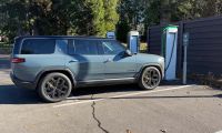 A silver 2025 Rivian R1S is shown from the rear three-quarter view at an EV charging station, featuring black accent wheels and trim.
