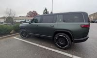 Forest Green 2025 Rivian R1S SUV photographed from a rear three-quarter angle in a parking lot, showing its boxy design, black wheels, and matte finish.