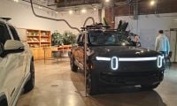 2025 Rivian R1S electric SUV in black, front three-quarter view, displaying distinctive LED headlight bar, shown in industrial-style showroom with exposed brick walls.