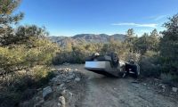 Tesla Cybertruck overturned on a narrow mountain dirt road surrounded by pine trees and rocky terrain