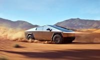 Tesla Cybertruck shown in side profile, kicking up dust while driving through desert terrain with mountains in background, showcasing its distinctive angular stainless steel design.