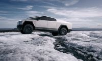 Tesla Cybertruck driving across icy, rugged terrain under a bright sky, showcasing its off-road performance and stainless-steel design.