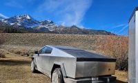 A Tesla Cybertruck in silver/stainless steel finish is parked in a mountainous landscape, shown from a rear three-quarter angle with snow-capped peaks visible in the background.