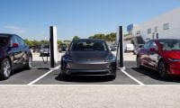 Multiple Tesla Model 3 vehicles in gray and red parked at charging stations, with white Supercharger pedestals visible, outside a modern commercial building.