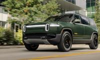 Green Rivian R1S electric SUV driving through a modern city street with glass office buildings in the background