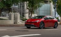 Red 2025 Toyota Prius driving through an urban street, showcasing sporty exterior styling and updated front fascia.