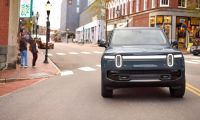 Front view of a Rivian R1S electric SUV driving through an urban downtown street, featuring signature vertical LED headlights and a boxy adventure-focused design.