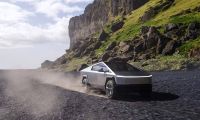 A silver Tesla Cybertruck kicking up dust while driving at speed on a gravel road along a dramatic, rocky coastline.