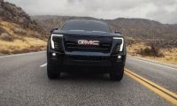 Black GMC pickup truck, front view on desert highway, featuring LED headlights, bold grille, rocky mountain landscape background.