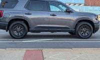 2026 Honda Passport TrailSport Elite in gray shown in full side profile parked on a city street with modern buildings in the background.