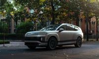 A silver Hyundai Ioniq 9, viewed from a front three-quarter angle, parked on a tree-lined historic city street with brick buildings in the background.
