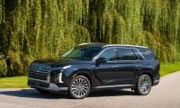 2026 Hyundai Palisade in black, photographed at three-quarter front angle on paved road beneath weeping willow trees, featuring distinctive split LED headlights and large grille.