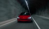 A red Tesla Model 3 shown from the front three-quarter angle driving through a dimly lit tunnel, with its distinctive LED headlights illuminated and creating a dramatic atmosphere.
