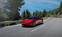 Candy red Tesla Model 3 driving on a winding mountain road, photographed from front three-quarter view, surrounded by pine trees and guardrails.