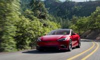 Front three-quarter view of a red 2026 Tesla Model S in motion on a curving road through a forested landscape