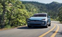 A light blue Tesla Model X SUV shown from a front three-quarter angle, driving on a winding mountain road surrounded by lush green forest.