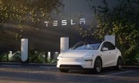 White Tesla Model Y parked at a Tesla Supercharger station with Tesla logo signage in the background.