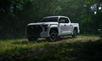 2026 Toyota Tundra in white, photographed at three-quarter front angle in a wooded setting, featuring black accents and distinctive alloy wheels.
