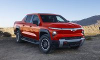 2026 Chevrolet Silverado EV electric pickup truck in red, front three-quarter view off-roading on desert terrain with rugged tires and bold grille design.