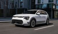 White 2026 Kia Niro EV driving through a city street in a front three-quarter action view.