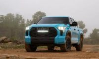 A head-on shot of a light blue 2025 Toyota Tundra TRD Pro with its distinctive LED light bar turned on, parked in a muddy, off-road environment.