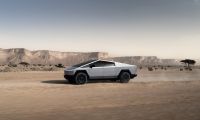 A silver Tesla Cybertruck shown in side profile view against a desert landscape with rocky cliffs and scattered trees in the background.
