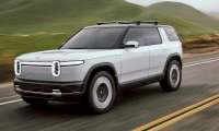 White 2027 Rivian R2 driving on a two-lane road through green hills in a front three-quarter action view.