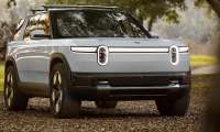 White 2027 Rivian R2 parked on a wooded trail in a front three-quarter view.
