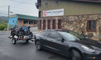 A black Tesla Model 3 is parked at a three-quarter front angle outside eMoto Powersports, with motorcycles on a trailer behind it.