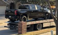 A black Chevrolet Silverado pickup truck being loaded onto a flatbed tow truck, photographed from the side angle in a residential driveway.