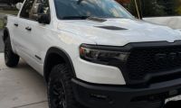 White 2025 Ram 1500 Rebel pickup truck shown featuring its distinctive wide body, hood scoop, and black trim accents.