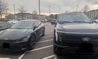 A black 2023 Kia EV6 and a dark blue Ford F-150 Lightning are parked side-by-side in a parking lot, both shown from their front three-quarter angles at dusk with apartment buildings visible in the background.