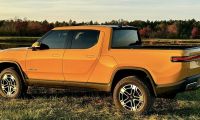 Orange Rivian R1S electric SUV shown from the rear three-quarter view, parked on grass near a lake with autumn trees in the background during sunset.