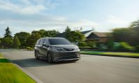 A dark gray 2024 Toyota Sienna minivan shown from the front three-quarter angle, driving on a suburban road with motion blur effect and green landscaping in the background.