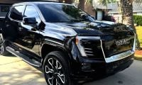 A black GMC Sierra EV shown from a front three-quarter angle parked in a residential driveway, featuring distinctive black and chrome wheels and LED lighting elements.