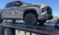 A tan 2025 Toyota Tundra with rugged tires is secured on a flatbed tow truck under a clear blue sky. The scene conveys a sense of utility and readiness.