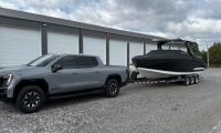A grey GMC Sierra EV parked on gravel in front of storage units, towing a large black boat with a triple-axle trailer. The scene is under a partly cloudy sky.
