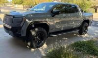 Gray GMC Sierra EV with black off-road wheels, lifted suspension, and custom front bumper parked on residential driveway in suburban setting.