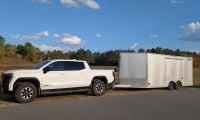 A white GMC Sierra EV is parked on a paved road, towing a large white enclosed trailer. The background features a grassy field and a clear, sunny sky.