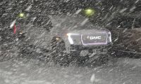 A GMC Sierra EV photographed during heavy snowfall at night, with its illuminated GMC badge visible through the swirling snow and yellow lights glowing in the background.
