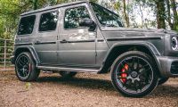 A gray Mercedes-Benz G-Class SUV shown from a side-front quarter view, featuring black aftermarket wheels, red brake calipers, and photographed in a wooded setting.