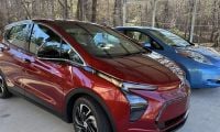 A maroon/red 2022 Chevrolet Bolt EUV is shown from a three-quarter front angle, parked next to a blue Nissan Leaf under a covered carport surrounded by trees.