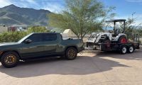 A black Rivian R1T electric pickup truck is shown from a side/front 3/4 angle, parked on a concrete pad with desert mountains visible in the background. The truck appears to be equipped with aftermarket bronze/gold wheels and a roof rack system.