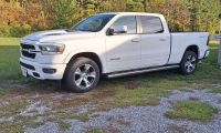 White 2024 Ram 1500 pickup truck shown from a front three-quarter view, parked on gravel with trees in the background, featuring chrome accents and alloy wheels.