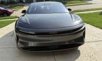 Sagar's 2024 grey Lucid Air Touring parked in a driveway, facing forward. The scene is suburban with neatly trimmed lawns and other cars in the background.