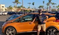 Scott wears a mask and is in casual clothing standing proudly beside an orange Chevy Bolt in a sunny parking lot filled with palm trees and parked vehicles.