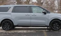 A grey Kia Carnival is shown from the side view, featuring black wheels and trim, photographed in a winter setting with snow-covered trees in the background.