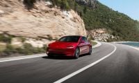 A red Tesla Model 3 speeds along a winding coastal road. Surrounded by rocky cliffs and greenery under a clear blue sky, the scene evokes freedom and adventure.