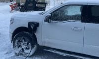 White Ford F-150 Lightning pickup truck shown from the side angle in snowy winter conditions, with snow-covered wheels and a utility vehicle with plow attachment visible in the background.